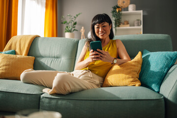 Asian woman relaxing on green couch while using smartphone, bright room