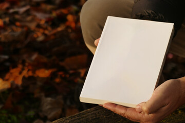 Mock up - man reading book with white cover in the park with autumn leaves and wooden background