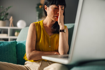 Asian woman in yellow top on green couch working on a laptop at home.