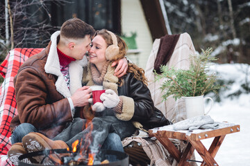 Portrait of pretty cheerful beautiful couple in warm clothes, knitted sweater embracing smiling having fun outdoors in winter in the park. Snowflakes, frosty day, snow all around. Countryside house