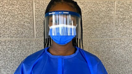 A woman wears a blue face mask and a clear face shield while standing in front of a brick wall