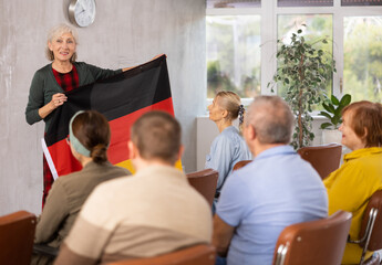 Fototapeta premium Positive friendly aged female tutor giving lecture to group of older adults during educational class, holding national flag of Germany and talking about history of country..