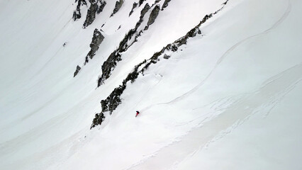 single snowboarder glides across a steep, open face of pure snow, framed by dark rocky outcrops.