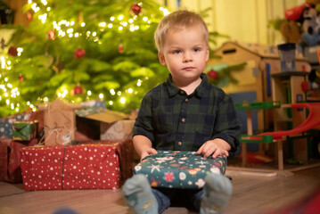 
two-year-old blond boy sits under the Christmas tree with gifts in his hands, is festively dressed in a shirt, carols are playing, Christmas lights are on
