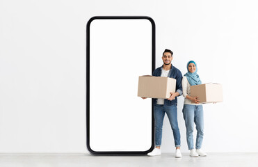 A young Muslim man and woman stand together, smiling while holding cardboard boxes. They are positioned next to a large empty smartphone display in a bright, minimalist space.