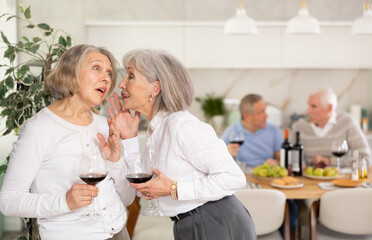 In cozy homely atmosphere, two elderly female friends have withdrawn from their male companions, stand with glass of wine in hands, communicate, discuss ladies secrets, share news