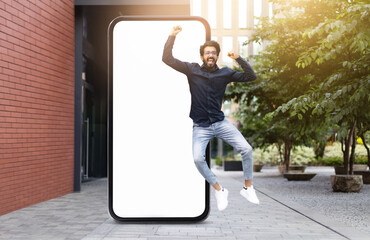 Indian man dressed in casual attire jumps joyfully in front of a large blank smartphone screen, situated in a sunny outdoor setting with greenery and modern architecture.