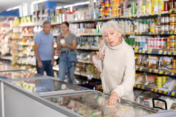 Positive interested gray-haired senior woman shopping in supermarket, choosing foods in glass display case