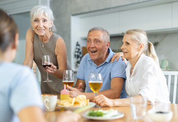 Group of cheerful elderly friends, women and man, gathering around kitchen table, enjoying beer and snacks, engaging in lively conversation and laughter