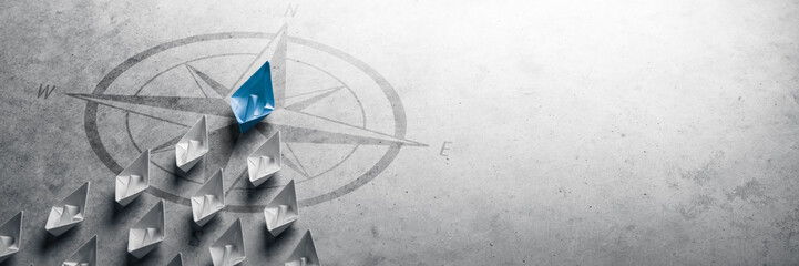 Blue Paper Boat Leading A Fleet Of Small White Boats With Compass Icon On Grey Concrete -...