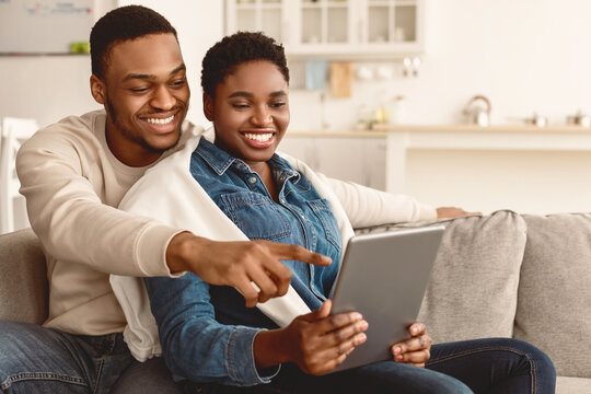 Happy loving young African American couple using digital tablet, watching video, looking at their photos or choosing what to buy, sitting and relaxing on couch in living room. Man pointing at screen