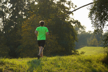 A man runs on a grassy path in a lush green park, wearing a neon green shirt and black shorts. A calm natural setting, the beauty and tranquility of exercising outdoors