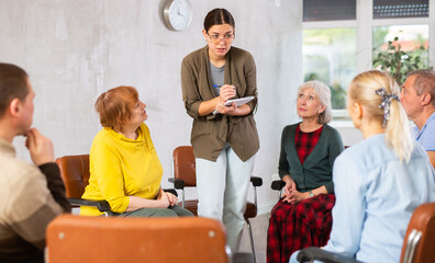 Friendly young girl conducting language speaking club, communicating with group of interested older...