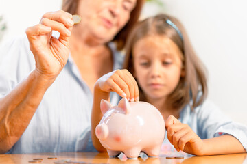 Grandmother and granddaughter inserting coins into piggy bank together, creating heartwarming...