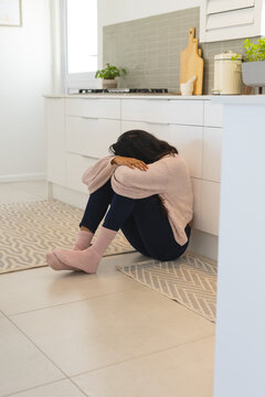 Young woman sitting on kitchen floor, feeling overwhelmed and contemplative, at home
