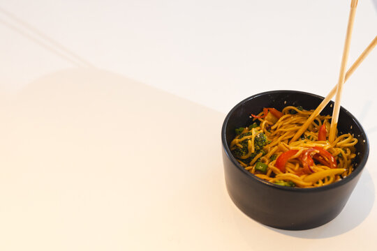 Chopsticks picking up colorful stir-fried noodles with vegetables in black bowl, copy space, at home