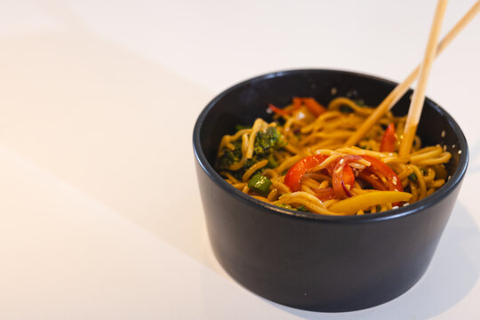 Chopsticks picking up stir-fried noodles with vegetables in black bowl, at home