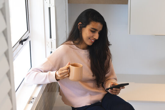 Relaxing at home, woman enjoying coffee and browsing smartphone by window