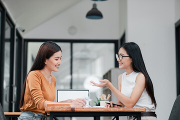 Two women smiling and talking at a desk in a modern office setting, showcasing teamwork and...