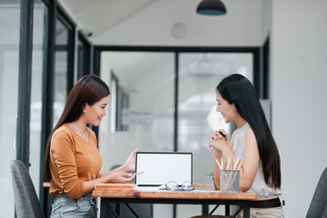 Two women in a modern office setting discussing business strategies with a laptop and tablet.