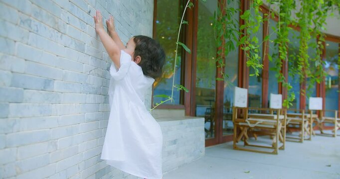 Cute child girl in a white dress Posing stands by a stone wall with Green Leaves, child exudes innocence and charm. adding a touch of nature to the serene
