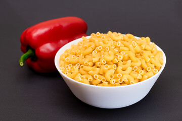 Uncooked Chifferi Rigati Pasta in White Cup and Red Bell Pepper on Black Background. Fat and Unhealthy Food. Scattered Classic Dry Macaroni. Italian Culture and Cuisine. Raw Pasta