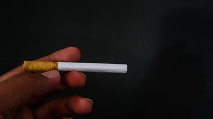 Close-up of a hand holding a cigarette against a dark background. The image highlights the cigarette's details, making it suitable for themes related to smoking, health, and lifestyle.