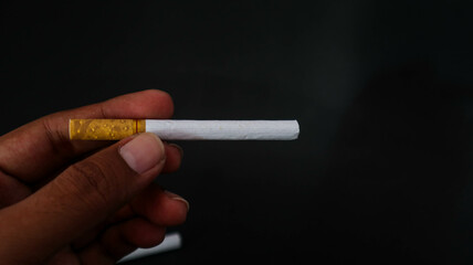 Close-up of a hand holding a cigarette against a dark background. The image highlights the cigarette's details, making it suitable for themes related to smoking, health, and lifestyle.