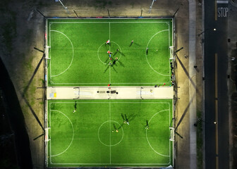 Aerial shot of a soccer field complex captured from directly above, with a drone. The image highlights two adjacent fields under bright floodlights, with players in action on both fields. The o © Visual Art Complex