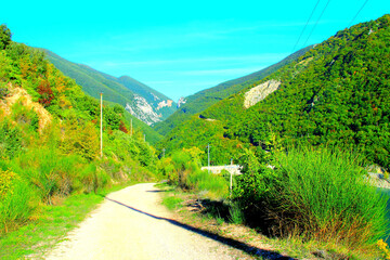 Wild scenery from Lago di Fiastra (Lake Fiastra) with a modest gravelly path stretching and winding between the green-topped Sibillini mountains and their abundant flora under an autumn cyan sky