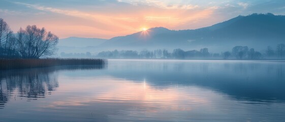Fototapeta premium Serene sunrise over a calm lake with mist, reflecting mountains and trees, creating a peaceful and tranquil natural landscape scene.
