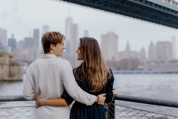 A couple shares a romantic moment by the tranquil water with a stunning city skyline in the background. New York.