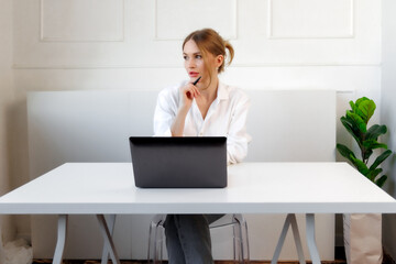 Young woman thoughtfully working on a laptop while sitting at a modern desk in a bright, minimalist...