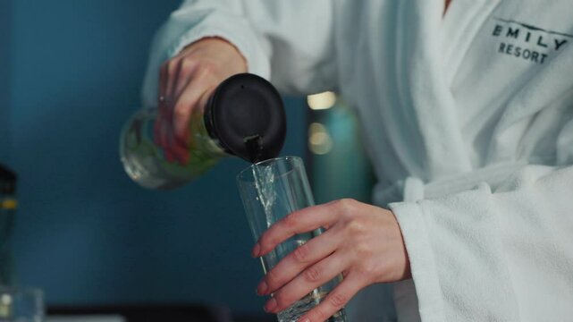 Footage of two unrecognizable women pouring water into glass. Female friends in white bathrobes drinking health drinks. Health program, diet at spa resort. Indoors
