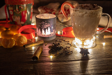 A mug of hot chocolate with whipped cream on top and a candy cane standing on a wooden rustic table. A candle is burning in a winter themed candleholder with traditional spices scattered around.