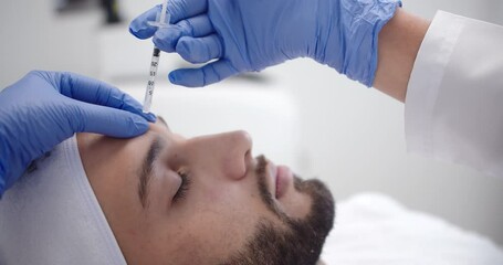 Beautiful slow motion shot of a female doctor's hand in blue gloves injecting botox into the forehead of a young bearded male patient lying on a stretcher.