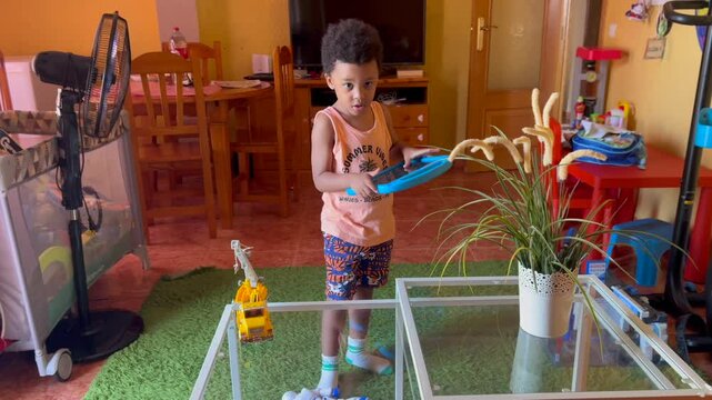4-year-old black kid playing with a tiny toy robot at home. He is wearing curly hair and summer clothes. Handheld camera. 4K