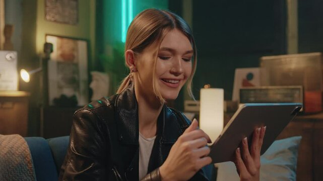 Shot of cute young stylish Caucasian woman in black leather jacket sitting on sofa and watching video on tablet. Attractive carefree female hipster touching screen of gadget in evening indoors