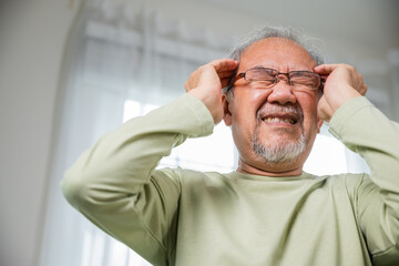 Headache. Close up of elderly holds head with hand suffering from migraine headache, Sad Asian senior man sitting on sofa feeling hurt and lonely at home, Old age health problems, healthcare