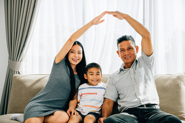 A cheerful family sits on a sofa parents gesturing a roof above their little son. Depicting themes of house insurance children protection and future planning ensuring family security.