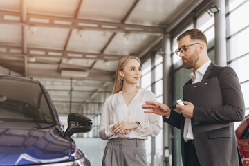 Car Sales Manager Showing Auto To Caucasian Lady Buyer Standing In Luxury Automobile Dealership Store. Buying Vehicle Concept