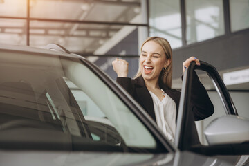 Cheerful woman standing near vehicle in dealership center and raising hands with excitement, copy...