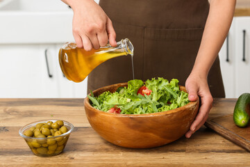 Woman adding olive oil into bowl with tasty salad at table in kitchen