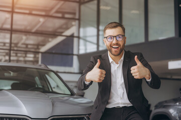 Bearded man standing near vehicle in dealership center and raising hands with excitement, copy space. Happy new owner male holding keys of new car after buying.