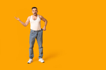 Fototapeta premium Handsome sporty shocked man in loose jeans on yellow background. Weight loss concept
