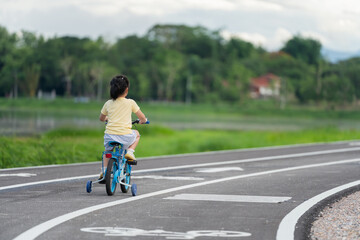 Obraz premium Asian young girl is riding a blue bicycle down a road. The road is lined with trees and there is a house in the background. The scene is peaceful and serene, with the girl enjoying her ride