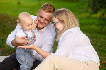 Family sitting on blanket on grass during summer walk in park. Happy child giving mother high five, clapping hand on palm. Family relationship concept, weekend, camping. Toddler playing with parents