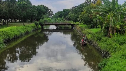 Fototapeta premium A pair of unrecognisable men in a small boat are fishing using electric equipment in the calm river, surrounded by lush greener