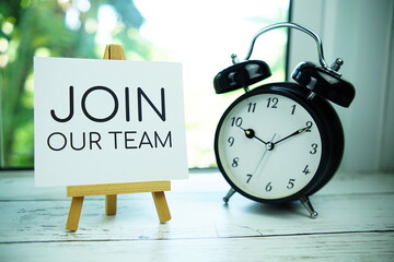 Join Our Team quotes text on paper card with easel and alarm clock in the window background