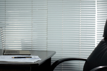 Modern wooden office desk with modern laptop computer, office chair and documents, blinds on background with copy space
